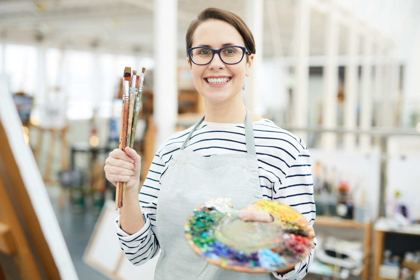An art therapist holds a paintbrush and palette and smiles at the camera.