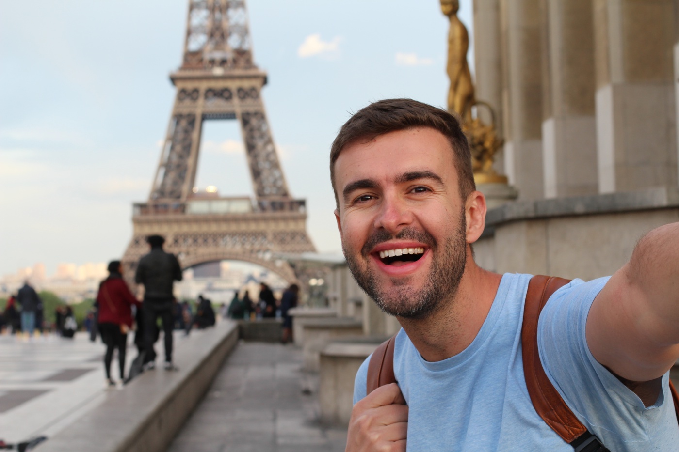 Ein Mann in einem Französisch Sprachaufenthalt steht vor dem Eiffelturm und lächelt in die Kamera.